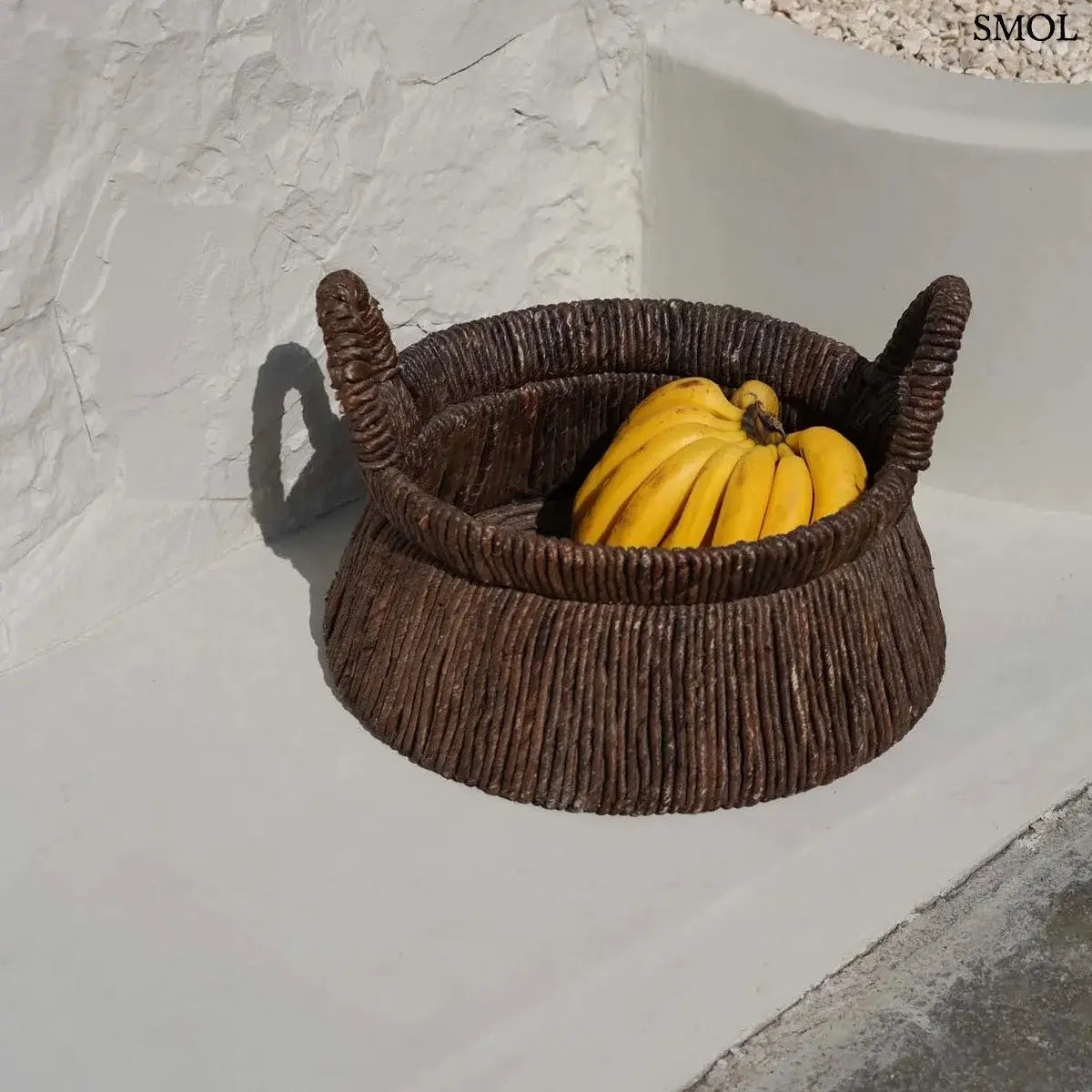 Kézzel font, sötétbarna banánlevél tárolókosár felfelé szűkülő háromszög alakú dizájnnal és masszív fogantyúkkal, teraszon