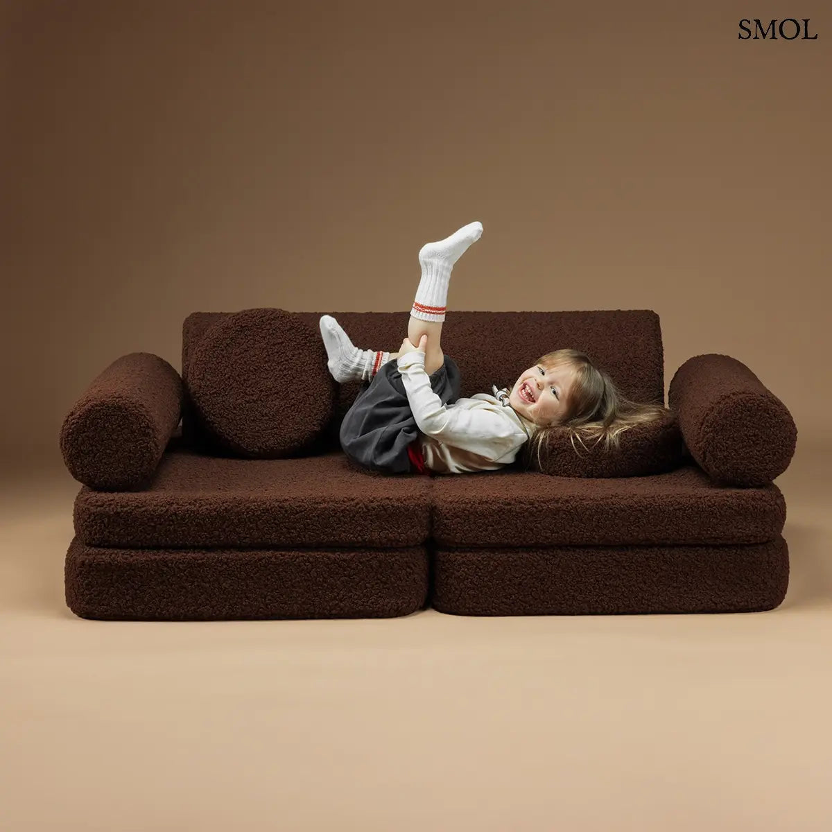 Barna, bársonyos tapintású kilencrészes gyerekkanapé henger és kerek párnákkal, puha teddy szövet borítással, barna háttérrel, rugdalózó gyerekkel