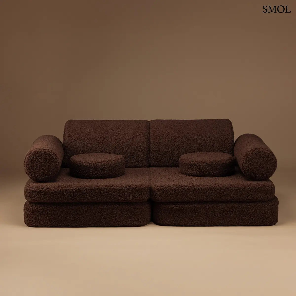 Barna, bársonyos tapintású kilencrészes gyerekkanapé henger és kerek párnákkal, puha teddy szövet borítással, barna háttérrel, szemből