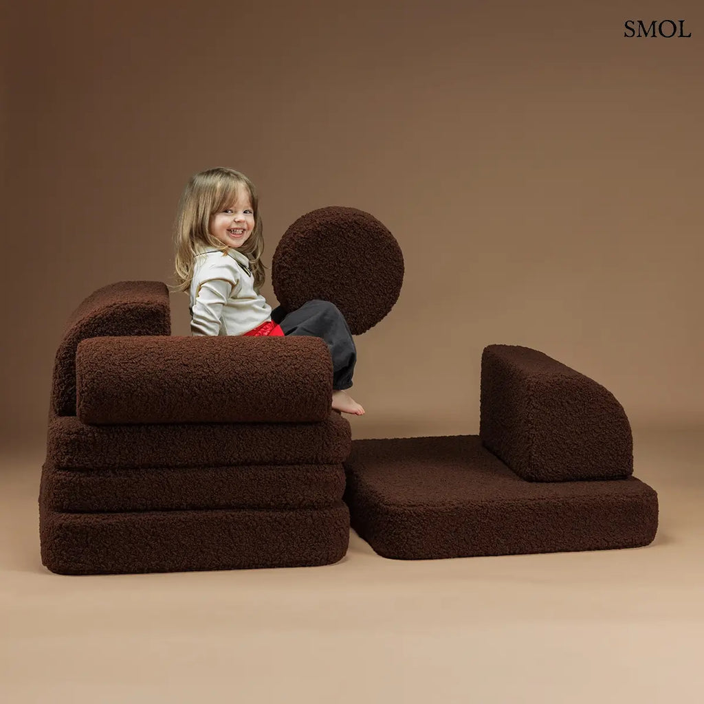 Barna, bársonyos tapintású kilencrészes gyerekkanapé henger és kerek párnákkal, puha teddy szövet borítással, elemekkel játszó gyerekkel, barna háttérrel