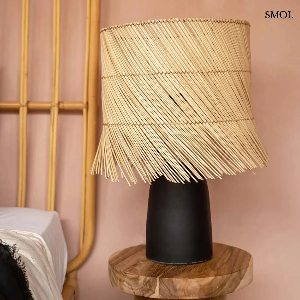 Modern asztali lámpa hengeres, fekete terrakotta talppal és natúr, ferdén szőtt rattan búrával, amelynek alsó széle játékosan rojtozott, hálószobában, ágy mellett
