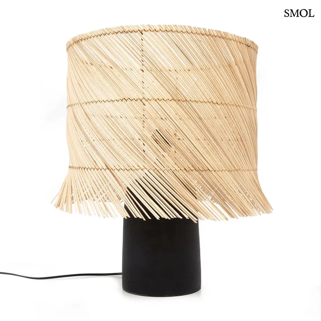 Modern asztali lámpa hengeres, fekete terrakotta talppal és natúr, ferdén szőtt rattan búrával, amelynek alsó széle játékosan rojtozott, fehér háttérrel
