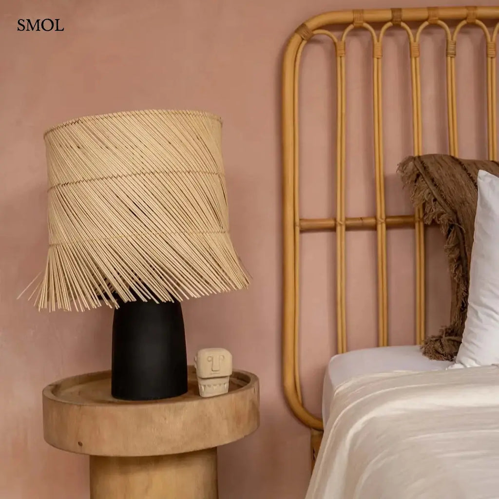 Modern asztali lámpa hengeres, fekete terrakotta talppal és natúr, ferdén szőtt rattan búrával, amelynek alsó széle játékosan rojtozott, hálószobában, szoborral