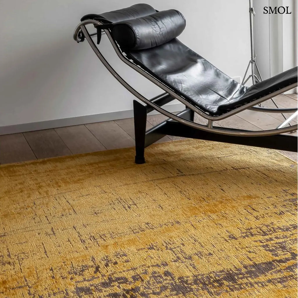 ECO RUG LIBERTY GOLD szőnyeg, aranyszín alapon sötétbarna és fekete festékszerű vonalakkal, action painting hatású mintázattal, fotellel nagyítva