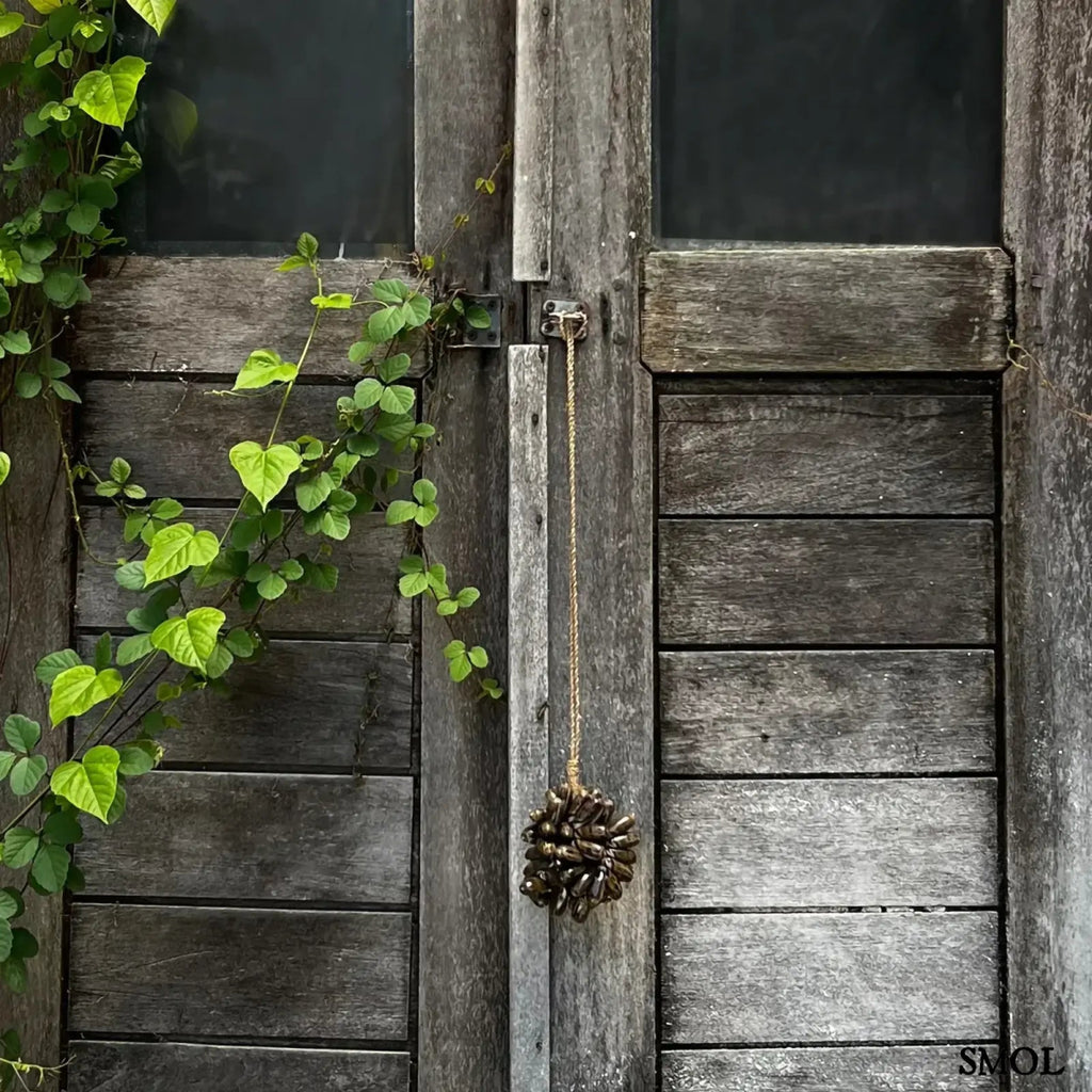  Barna kagyló függődísz, kézműves bojt formában, natúr kötélre akasztva szürke ajtón