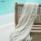 Kézzel szőtt, krémfehér pamut pléd gofri struktúrával és rojtokkal díszítve, medencével háttérben, fotel háttámláján