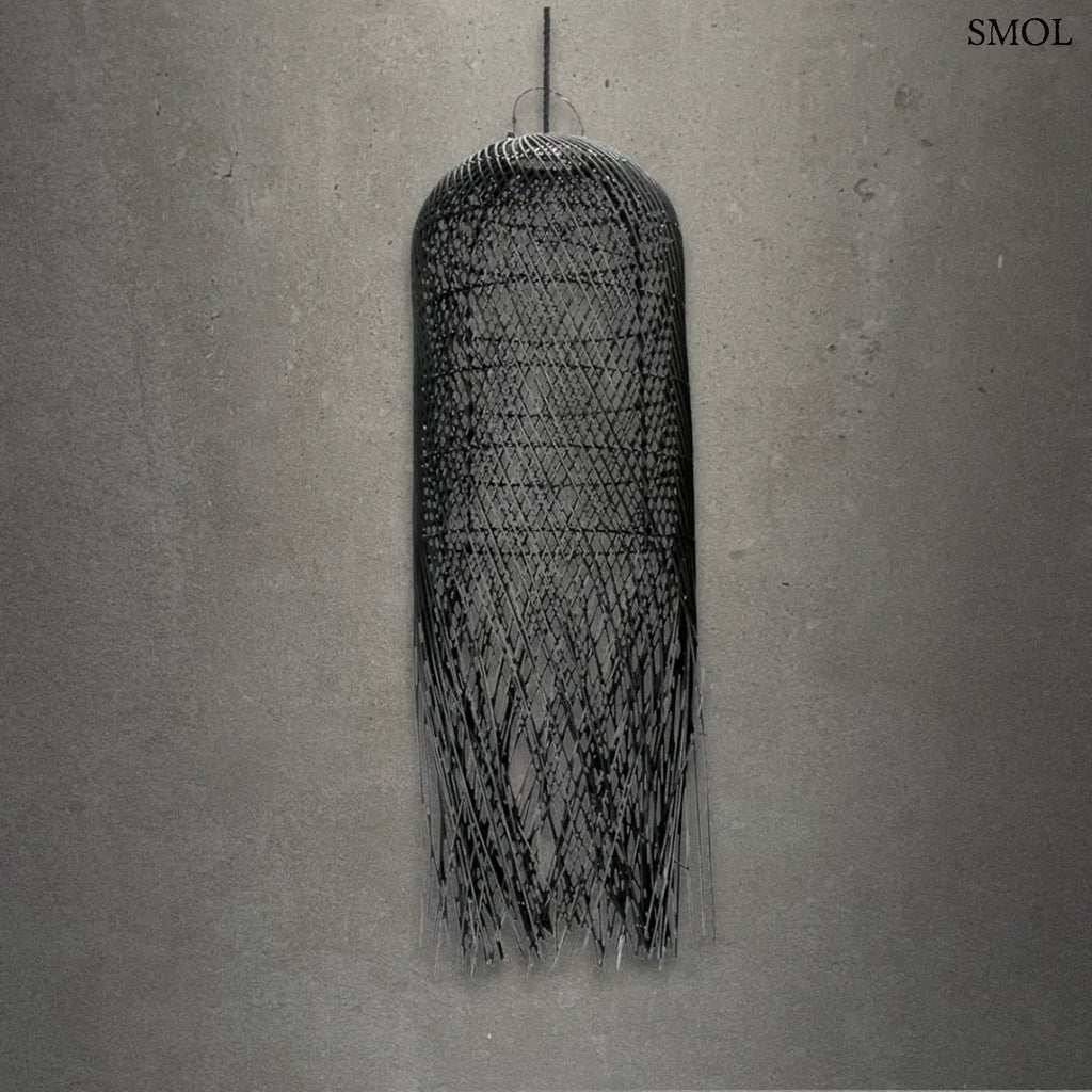 Hosszúkás, fekete színű, kézzel szőtt rattan lámpaernyő, amelynek alján a szálak szabadon, ritmikus vonalakkal végződnek, szürke háttérrel