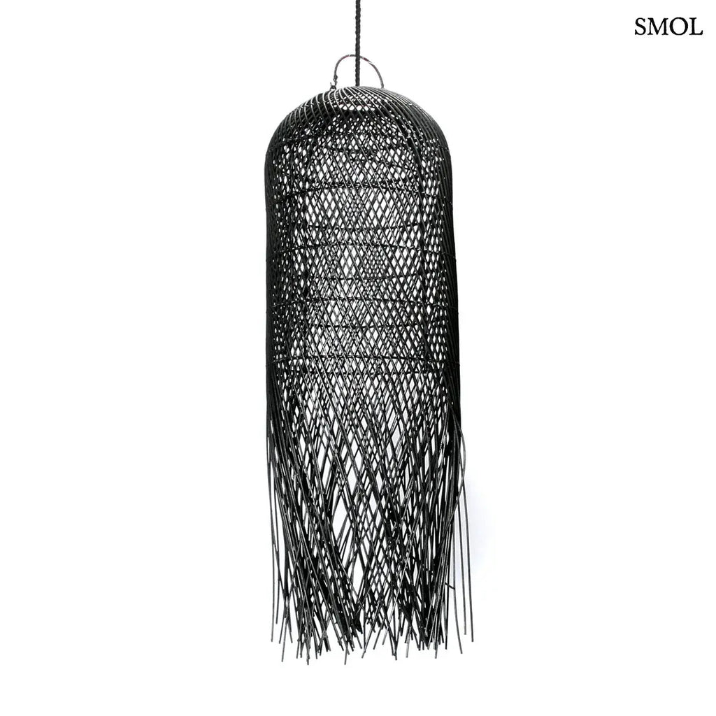 Hosszúkás, fekete színű, kézzel szőtt rattan lámpaernyő, amelynek alján a szálak szabadon, ritmikus vonalakkal végződnek, fehér háttérrel