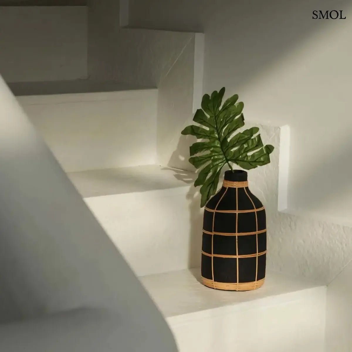 Terracotta váza fekete színben, natúr rattan fonással díszítve, szárazvirágokhoz, lépcsőn, levéllel