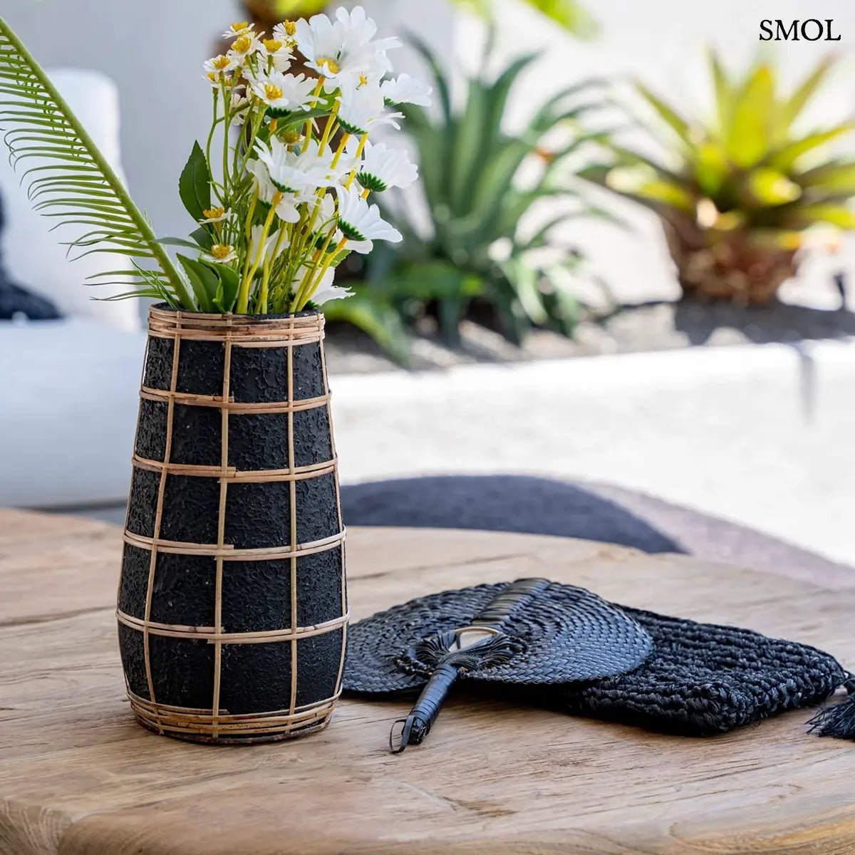 Fekete színű, rusztikus felületű terrakotta váza, amelyet világosbarna, kézzel font rattan rácsozat díszít, virágokkal
