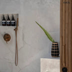 Fekete színű, rusztikus felületű terrakotta váza, amelyet világosbarna, kézzel font rattan rácsozat díszít, fürdőszobában