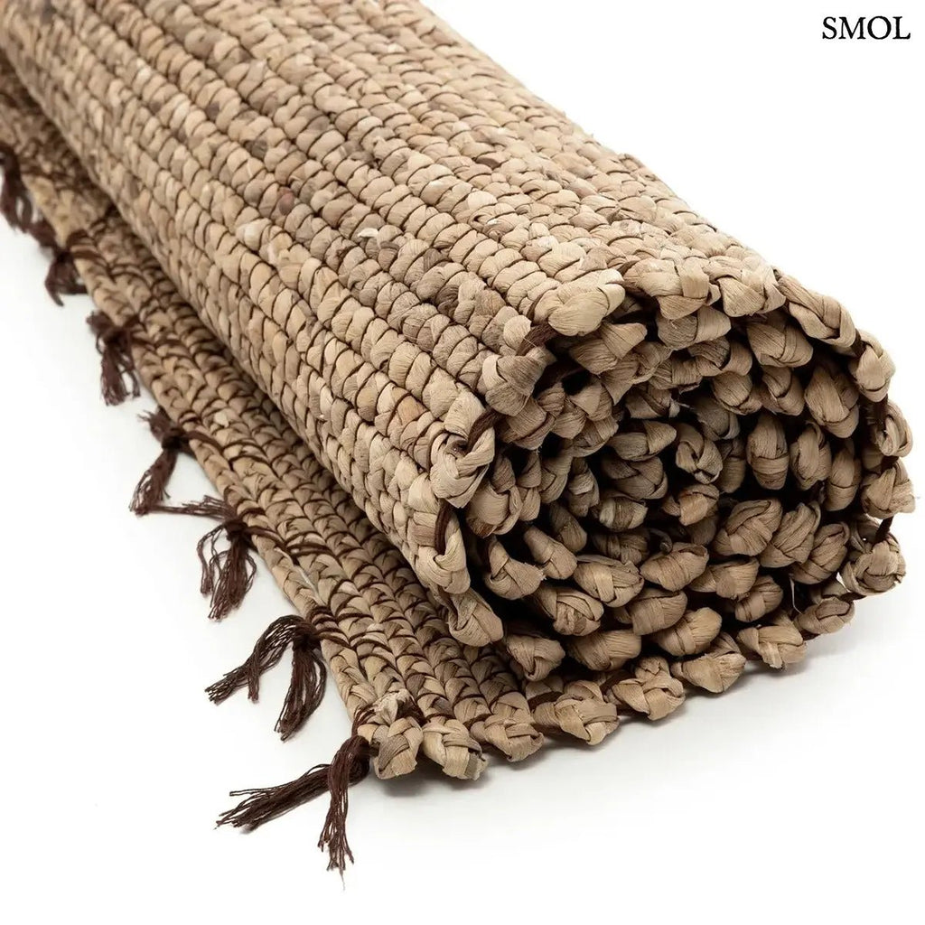 Kézzel szőtt, világosbarna vízijácint szőnyeg, jellegzetes fonott textúrával és sötétebb, csomózott rojtos szegéllyel, feltekerve